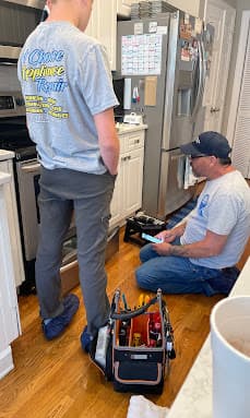 Jim kneels by a refrigerator with a tablet while his son Alex stands beside him on a service call