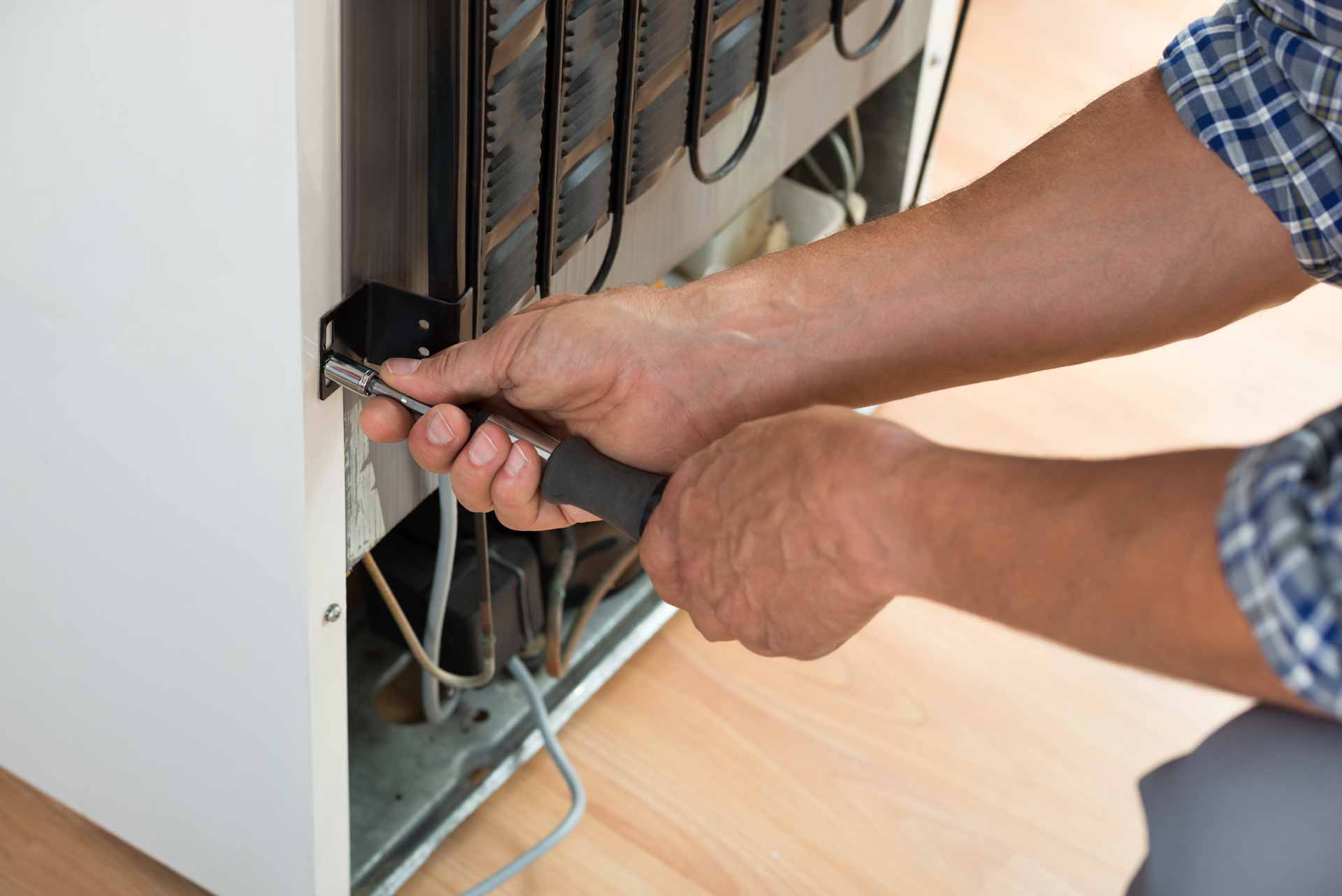 Technician tightening a fitting on the back of a refrigerator beside the condenser coils